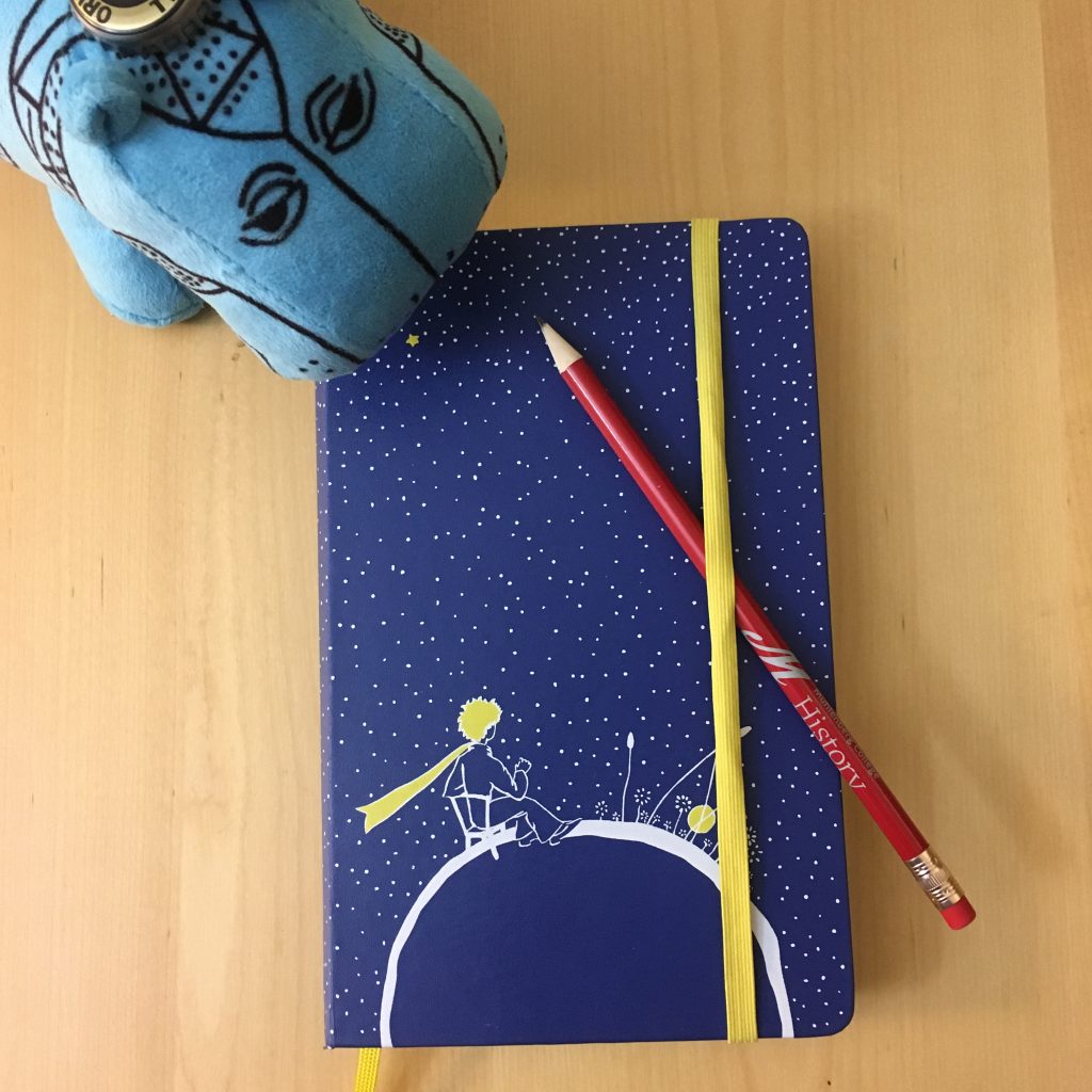 Blue planner with yellow and white picture of the Little Prince (Petit Prince) on cover, History Department Muhlenberg branded pencil under yellow elastic band; top left corner William the turquoise plush hippo