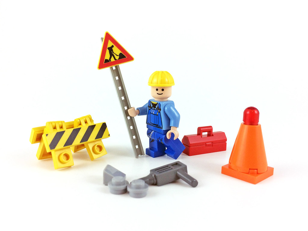 lego man and accessories: he has a hard hat, holds a  roadworks sign on a pole, and is surrounded by traffic cone, barricade, and tools